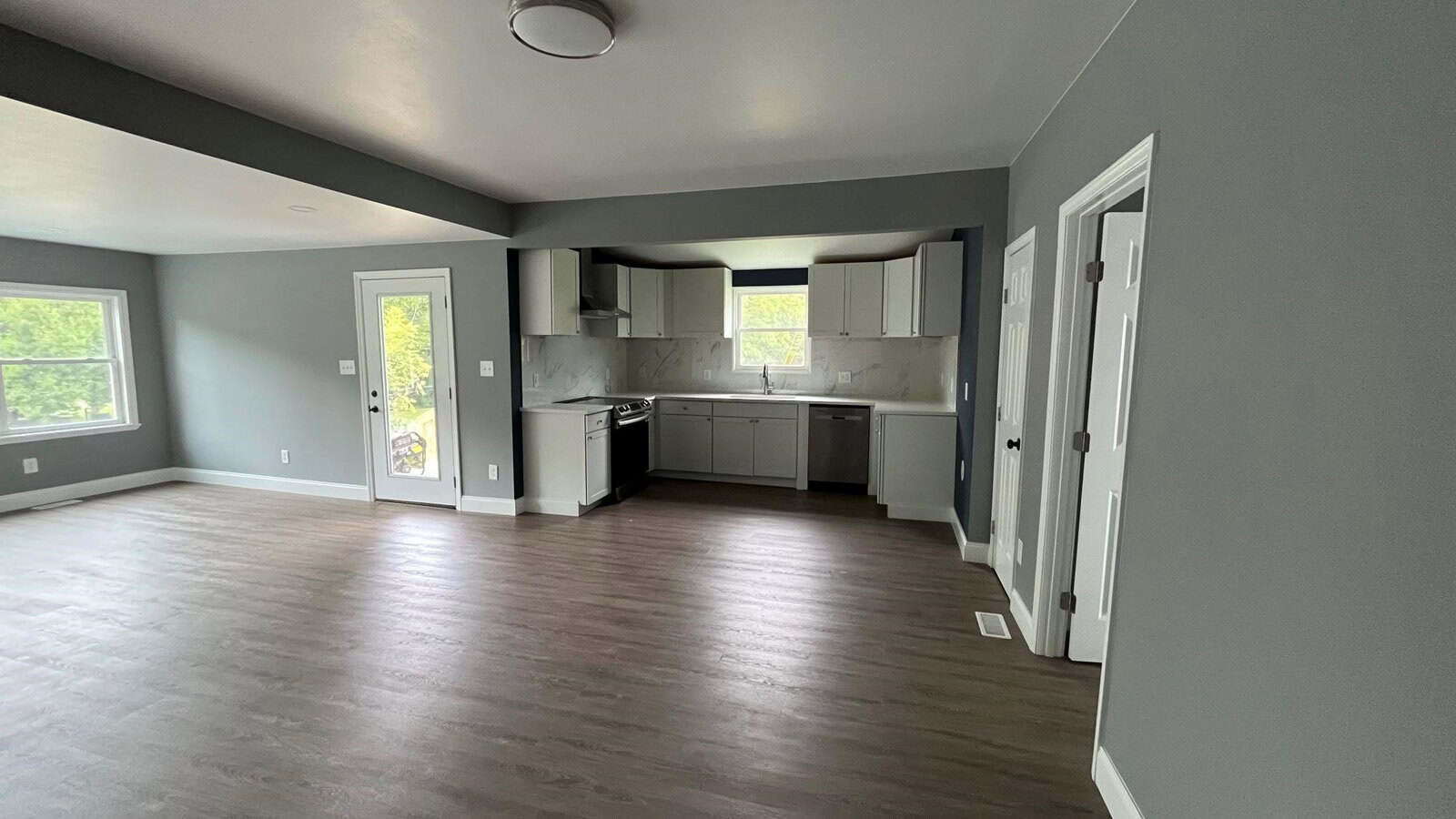 A modern kitchen and living area with gray walls and wood flooring. The kitchen features white cabinets, a stove, and a refrigerator. Large windows let in natural light, and there's a glass door leading outside. The space is open and unfurnished.