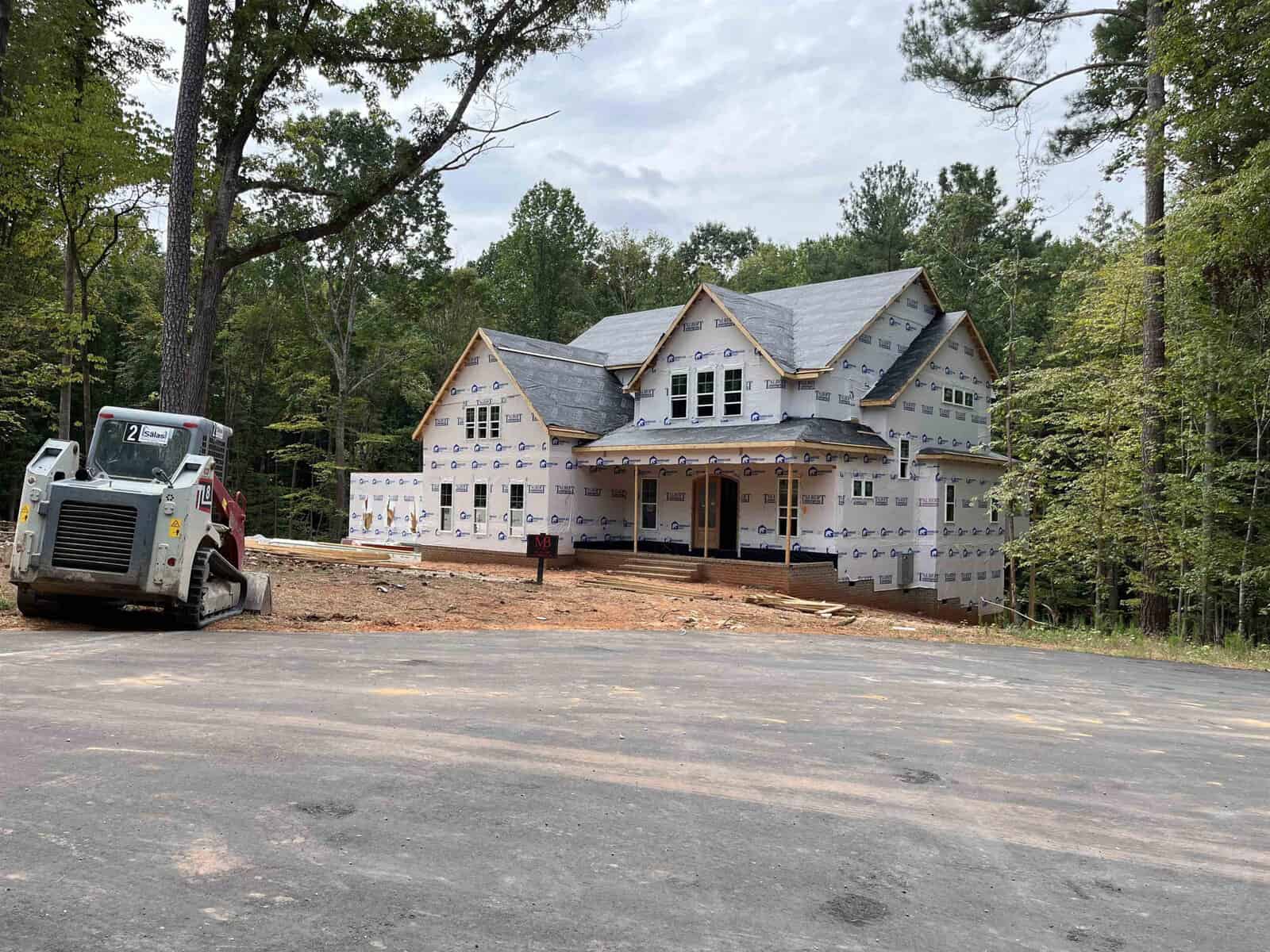A two-story house under construction is surrounded by trees. The exterior is partly covered with weather-resistant sheeting. A skid steer loader is parked on a dirt area in front of the house, with a paved driveway in the foreground.