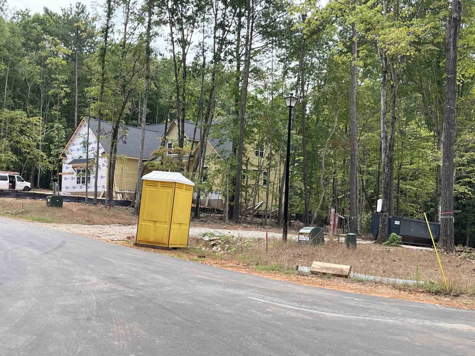 A house under construction surrounded by trees, with a yellow portable toilet in the foreground and a paved road curving to the left. Construction materials are visible around the site.