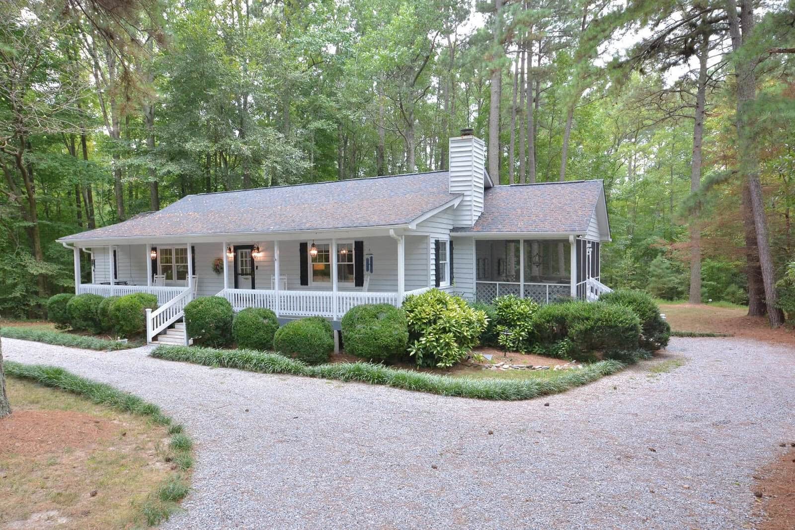 A single-story house with a gray gabled roof features a covered front porch, surrounded by green shrubbery and trees, with a gravel driveway leading up to it.