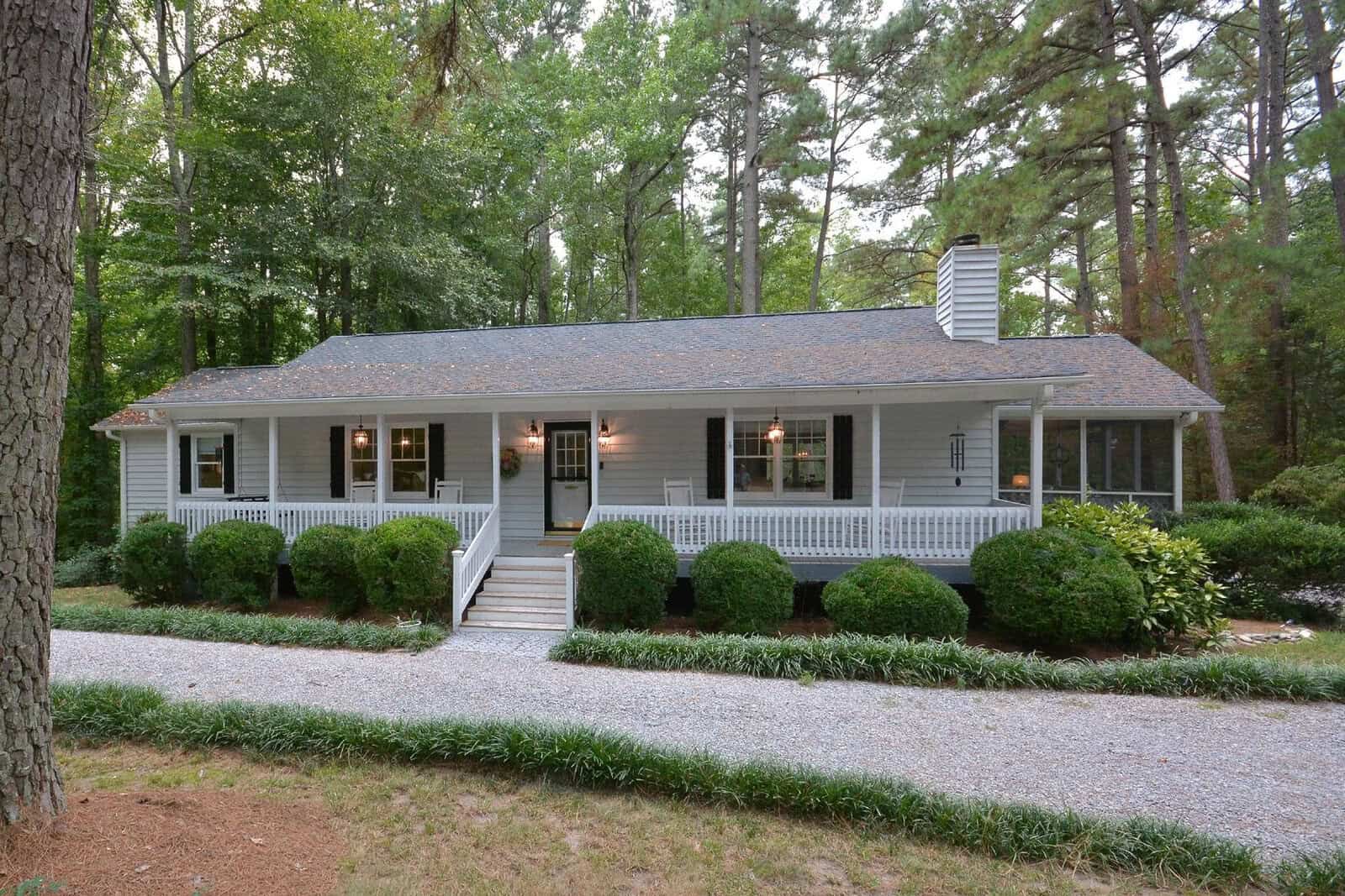 A single-story house with a gray roof and white siding, featuring a covered front porch with steps and surrounded by trees and a gravel driveway.