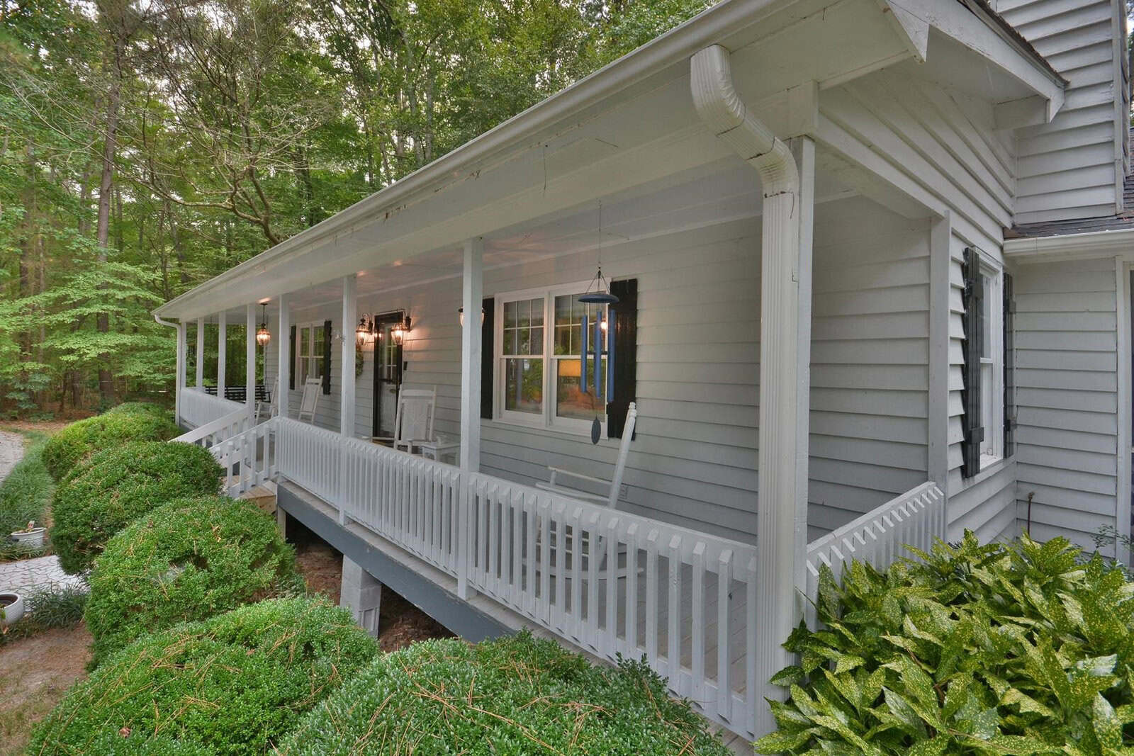 A side view of a gray wooden house with a covered porch, white railings, and landscaped bushes in a wooded setting.