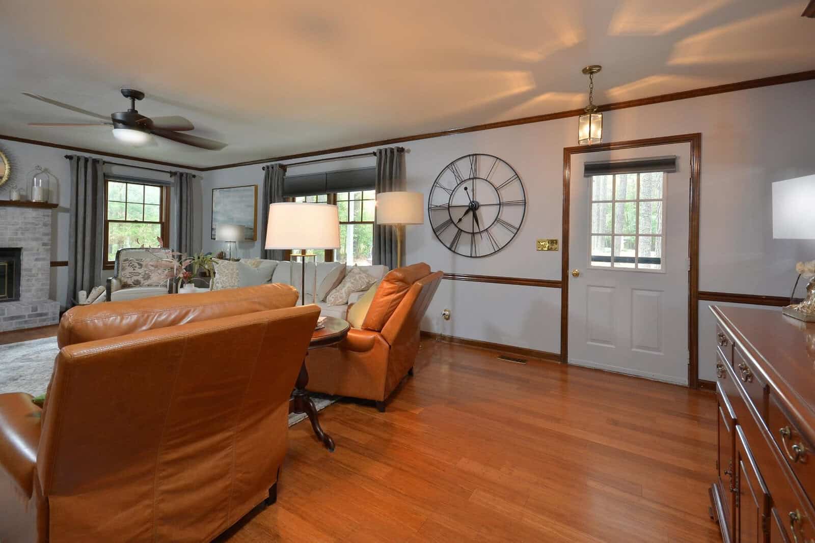 A spacious living room with hardwood floors, tan walls, a large wall clock, a leather armchair, and a white door.