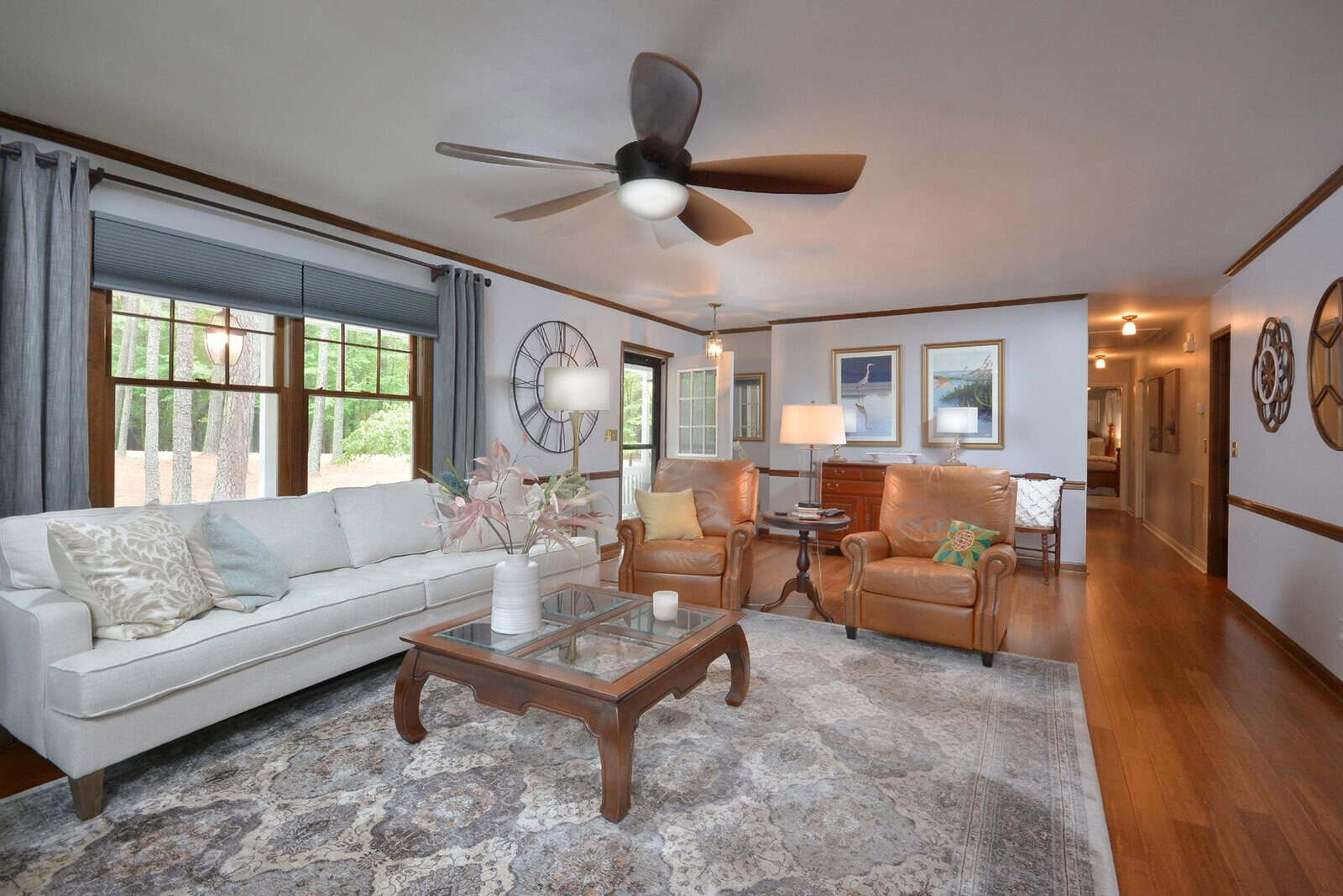 A well-lit, spacious living room with a combination of modern and traditional decor, featuring a beige sofa, two tan armchairs, a wooden coffee table, and hardwood flooring. the room is complemented by gray drapes, decorative items on the tables, and a ceiling fan above.