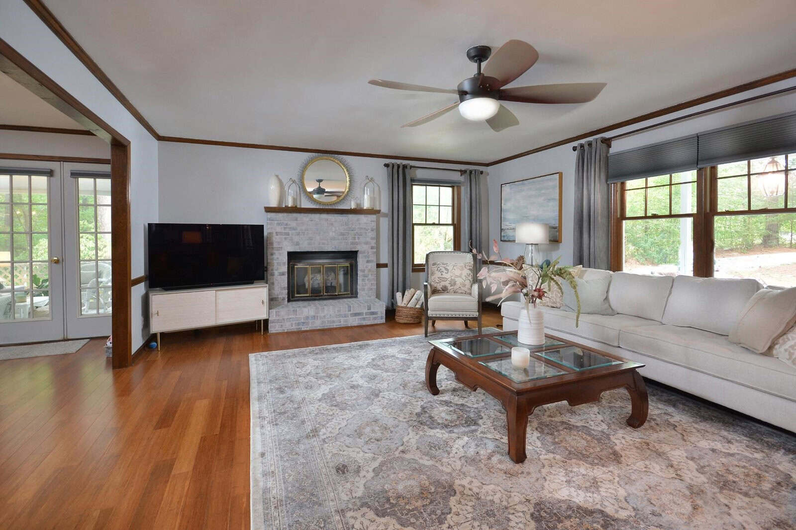 A spacious living room with hardwood floors, a sectional sofa, a fireplace, a ceiling fan, and windows providing natural light.