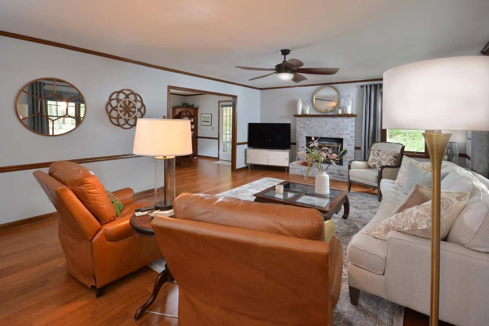 A well-lit, spacious living room with a mix of modern and traditional decor, featuring hardwood floors, a plush sofa, leather armchairs, a glass-top coffee table, and decorative mirrors on the wall.