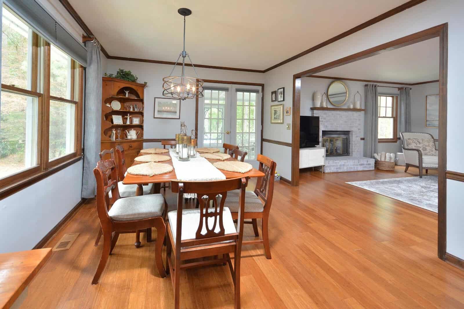 A spacious dining room featuring a wooden table set for six, with a hanging chandelier above it, adjacent to a living area with a fireplace and seating furniture. the room has large windows and is well-lit with natural light.