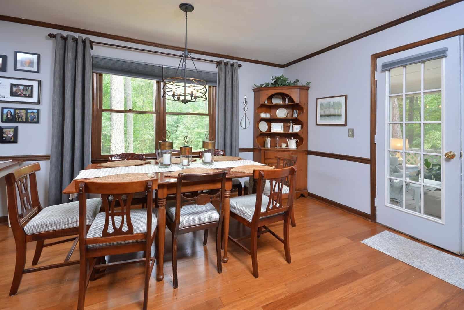 A traditional dining room with a wooden table set for a meal, matching chairs, hardwood flooring, and a hutch against the wall. the room features a window with greenery outside and a glass-paneled door leading to another area.