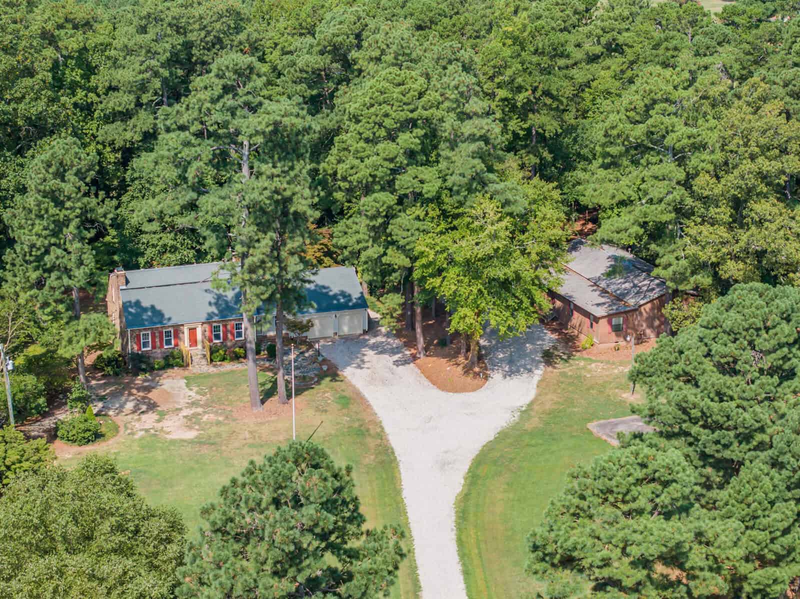 Aerial view of two residential buildings with red roofs surrounded by a large number of trees, featuring a winding driveway connecting them to a rural road.