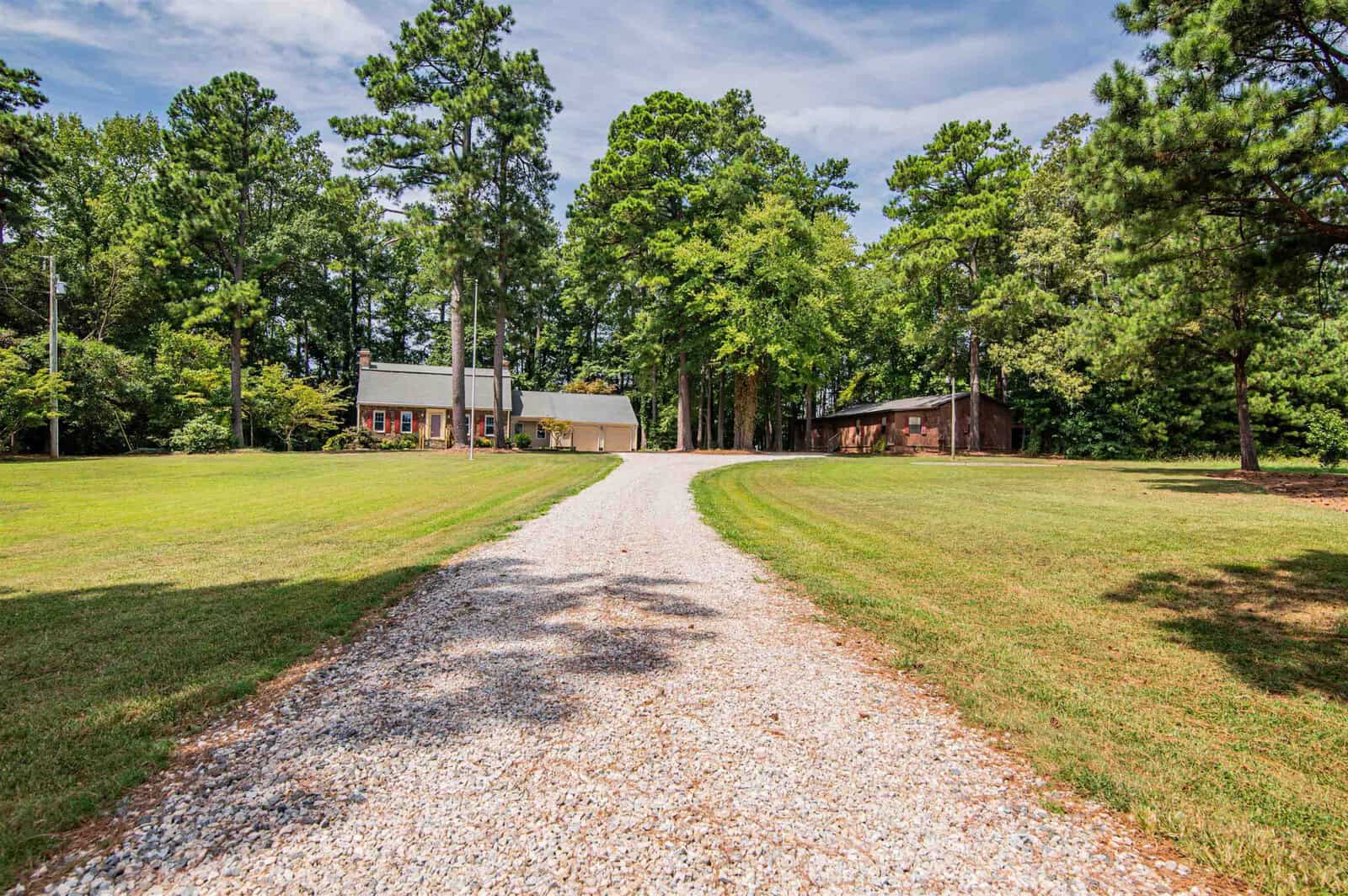 A gravel driveway leading to a house with a red brick exterior, surrounded by tall pine trees and a well-manicured lawn under a clear blue sky.