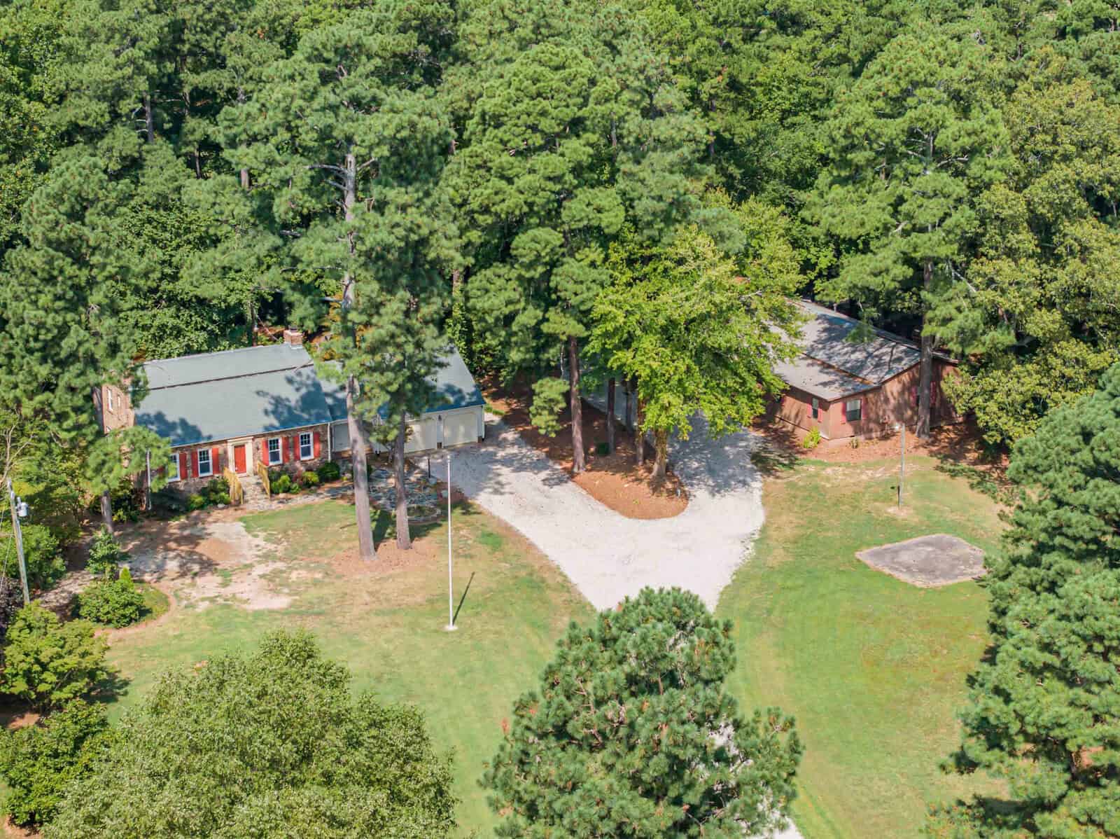 Aerial view of a single-story house with a red roof surrounded by green trees and a winding driveway.