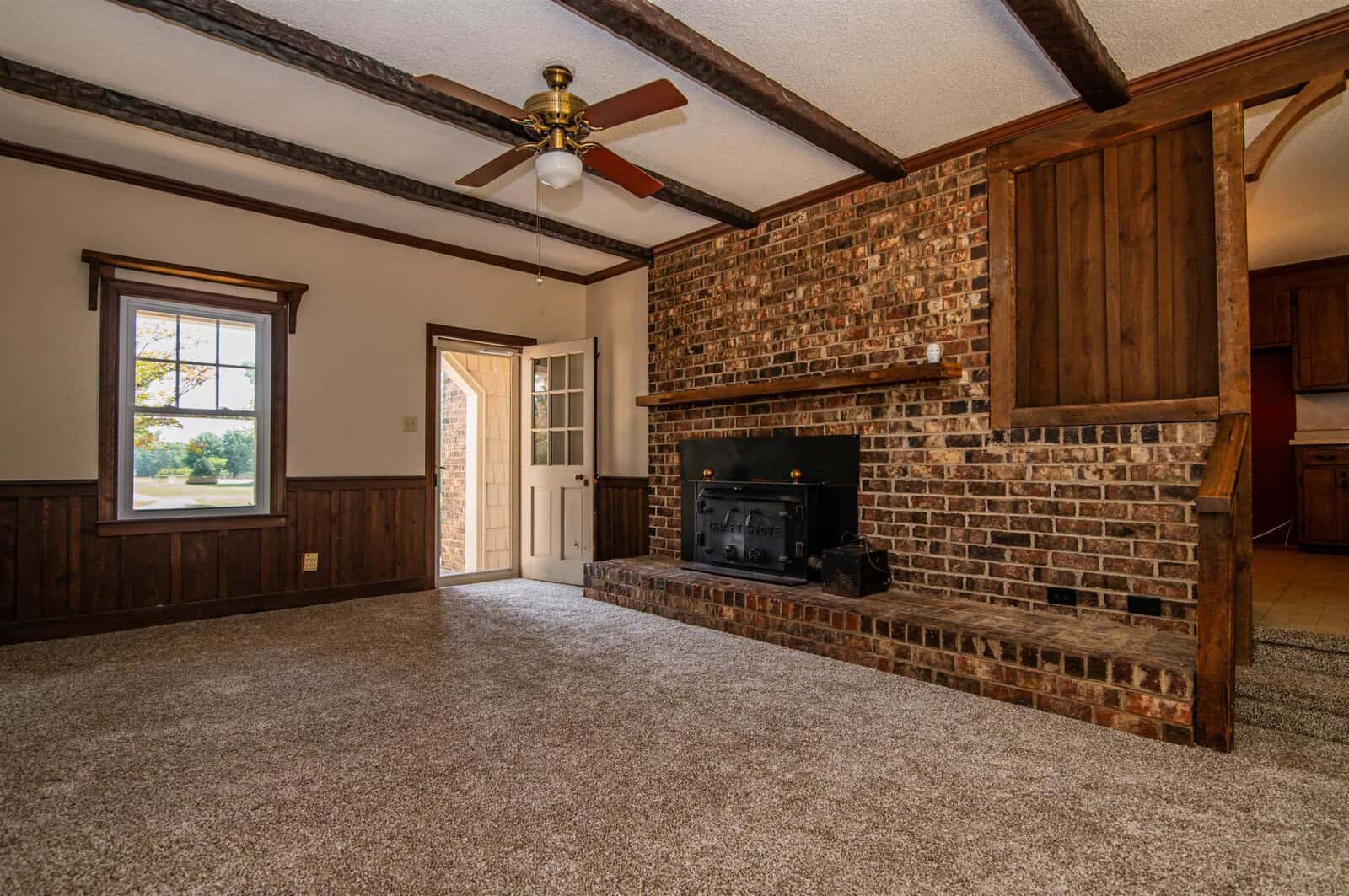 An empty room featuring a brick fireplace, wood paneling, and a carpeted floor with a staircase and exterior door visible in the background.
