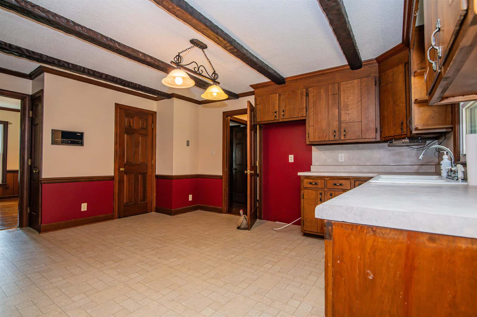 An empty kitchen featuring wooden cabinetry, a white countertop, and a beige tiled floor, with a visible dining area in the background.