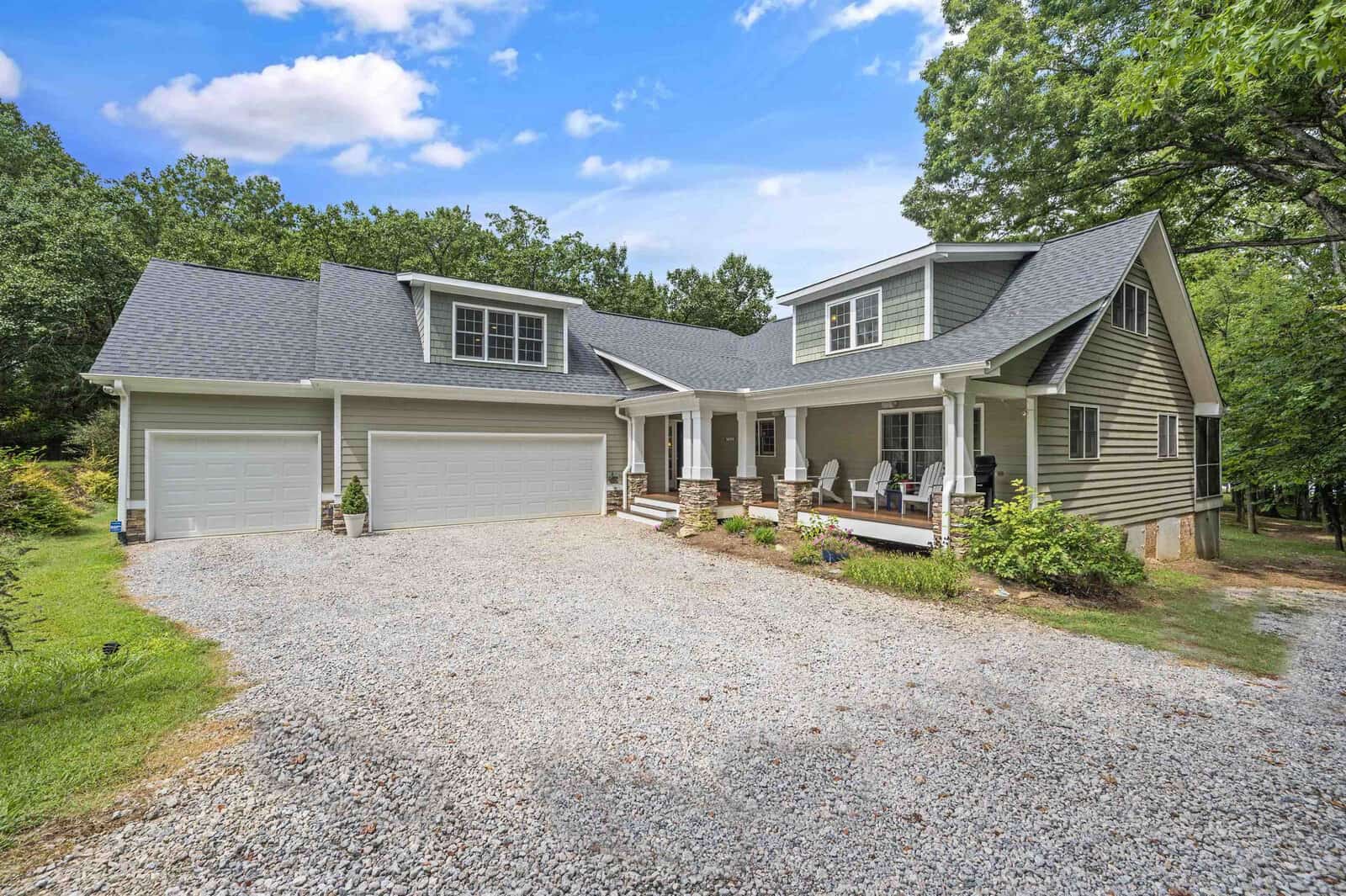 A detached single-family home with a beige exterior and a gray shingle roof features a two-car garage, a gravel driveway, and a front porch with white railings and outdoor seating, surrounded by trees under a clear sky.