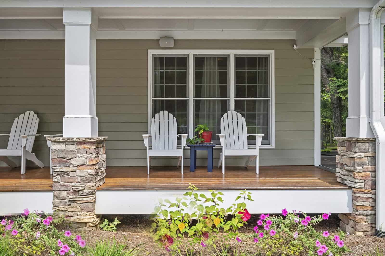 A cozy front porch with two white adirondack chairs flanking a small table with a red flowerpot, set against a home with gray siding and stone pillars, complemented by pink flowers in the foreground.