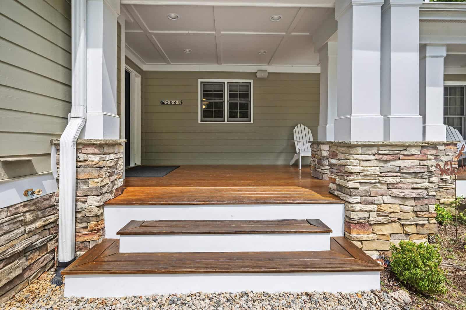 A neatly maintained entrance of a house featuring a wooden porch with steps, white columns, and stone cladding on the sides.