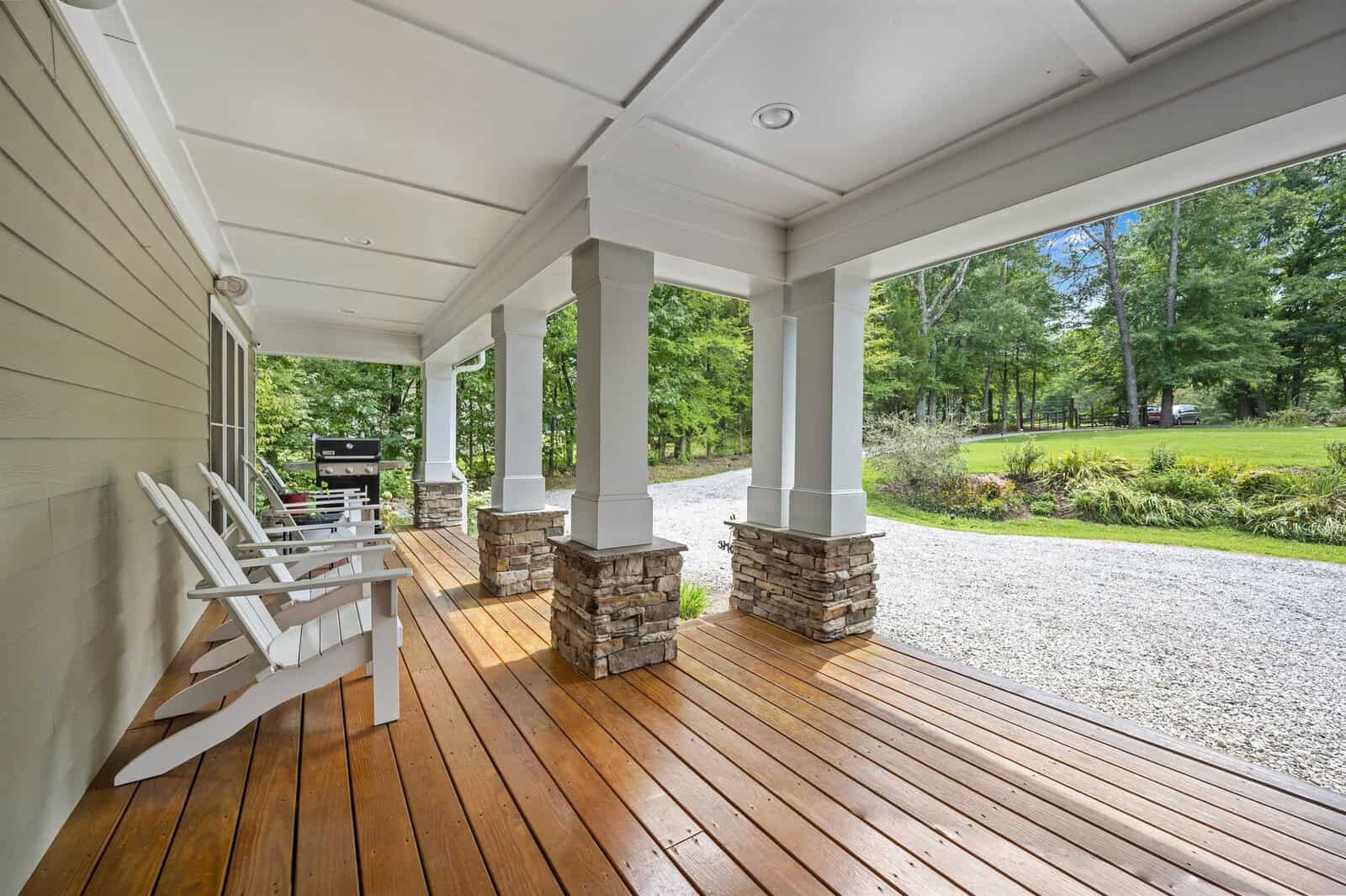 A spacious covered porch with white rocking chairs, wooden flooring, and stone-based columns overlooking a lush green yard.