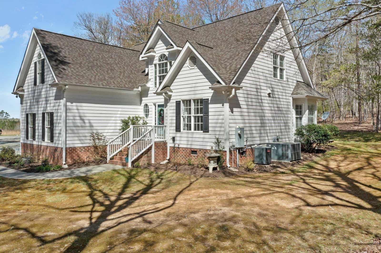 A two-story white house with a brick foundation, surrounded by trees. It features multiple gables, large windows, and a small porch with steps leading to the front door. Bare tree branches cast shadows on the lawn.