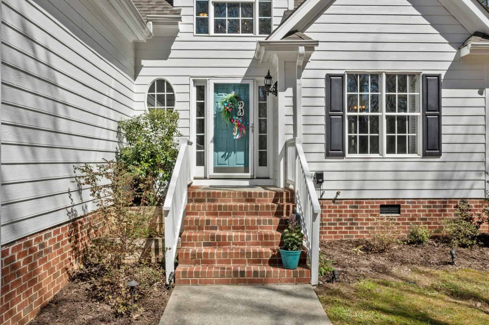 Steps lead to a bright blue front door with a colorful wreath on a white house. The house features brick accents, black shutters, and a few shrubs near the entrance. A paved walkway approaches the steps.
