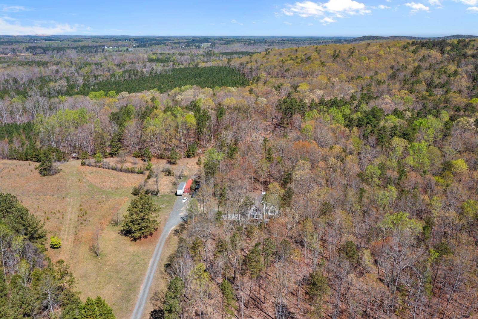 Aerial view of a rural landscape with a winding road leading to a house surrounded by dense forest. The area features a mix of green and brown trees, with clear skies above. Farmland is visible in the distance.