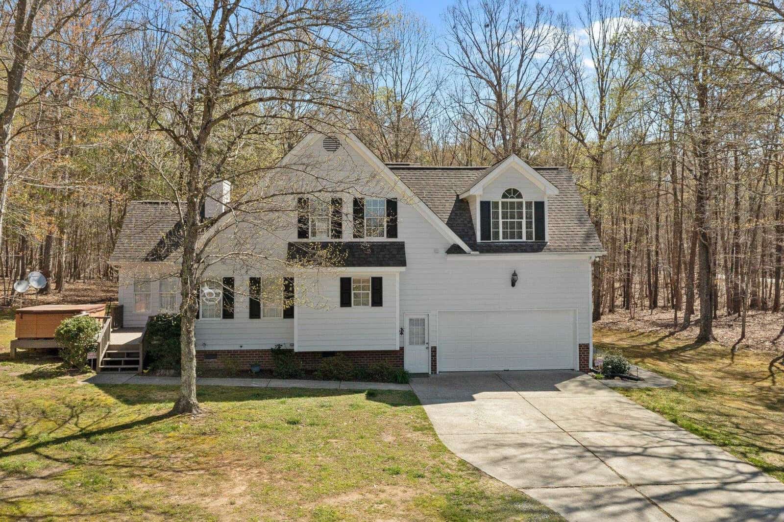 A two-story white house with black shutters, a double garage, and a gabled roof, surrounded by tall trees. The front yard has a large driveway and sparse grass. The house is set in a wooded area under a clear blue sky.