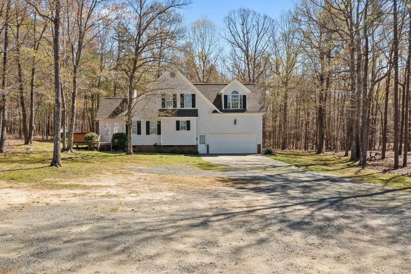 A white two-story house with black shutters, nestled among leafless trees. The house features a large front-facing garage and a gravel driveway. It's a sunny day, and the surrounding area is mostly wooded.