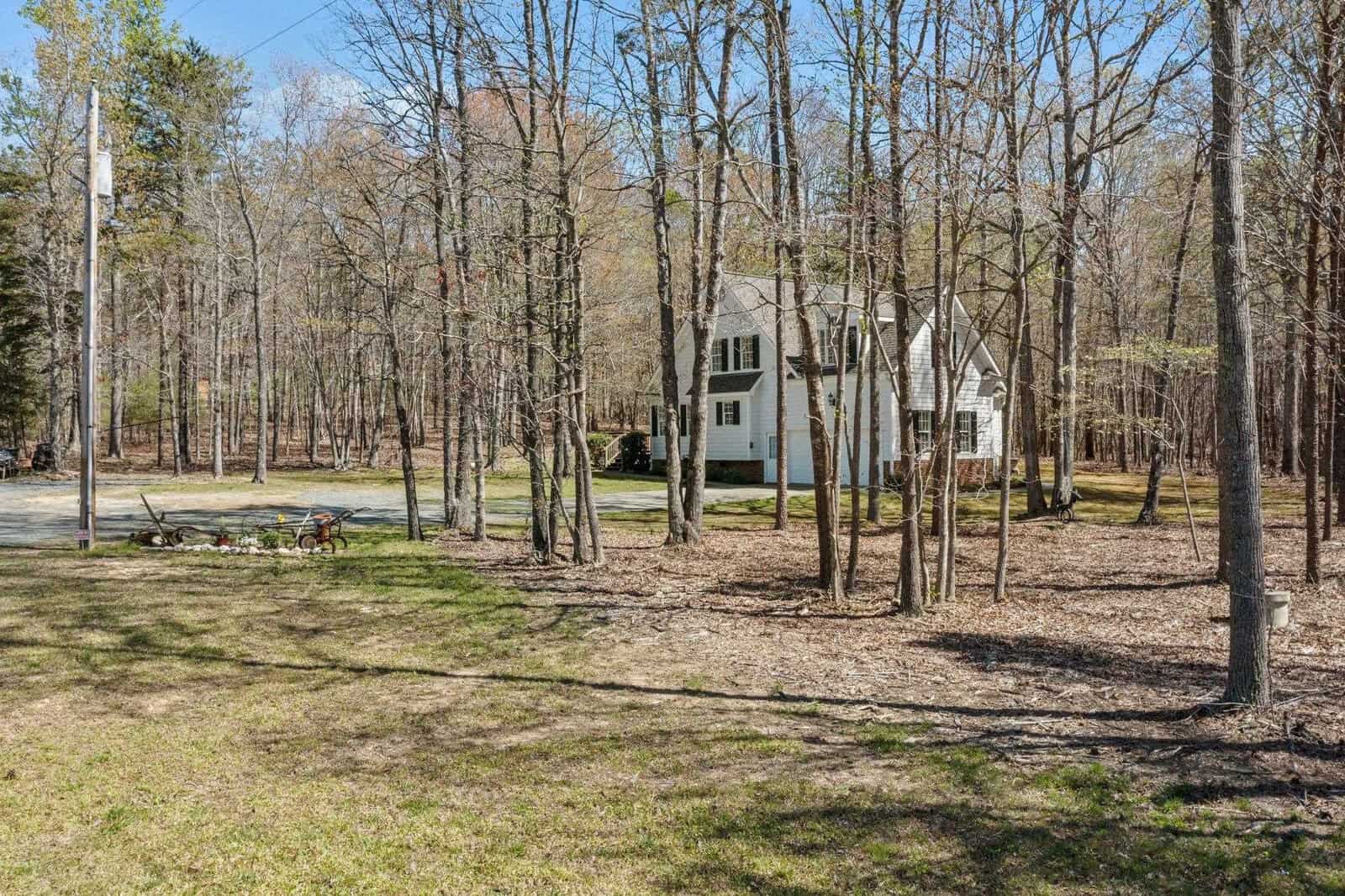 A white two-story house is surrounded by tall, leafless trees in a rural wooded area. A gravel driveway leads to the house, and there's a picnic table nearby. The sky is clear and blue, suggesting it's a sunny day.