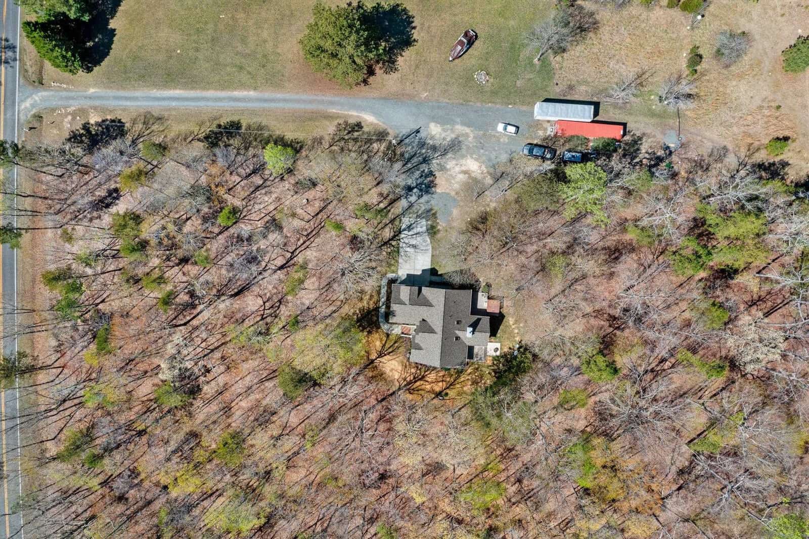 Aerial view of a house surrounded by trees, with a driveway leading to it. There are several vehicles parked nearby and a trailer visible. The surrounding landscape features a mix of grassy and wooded areas.
