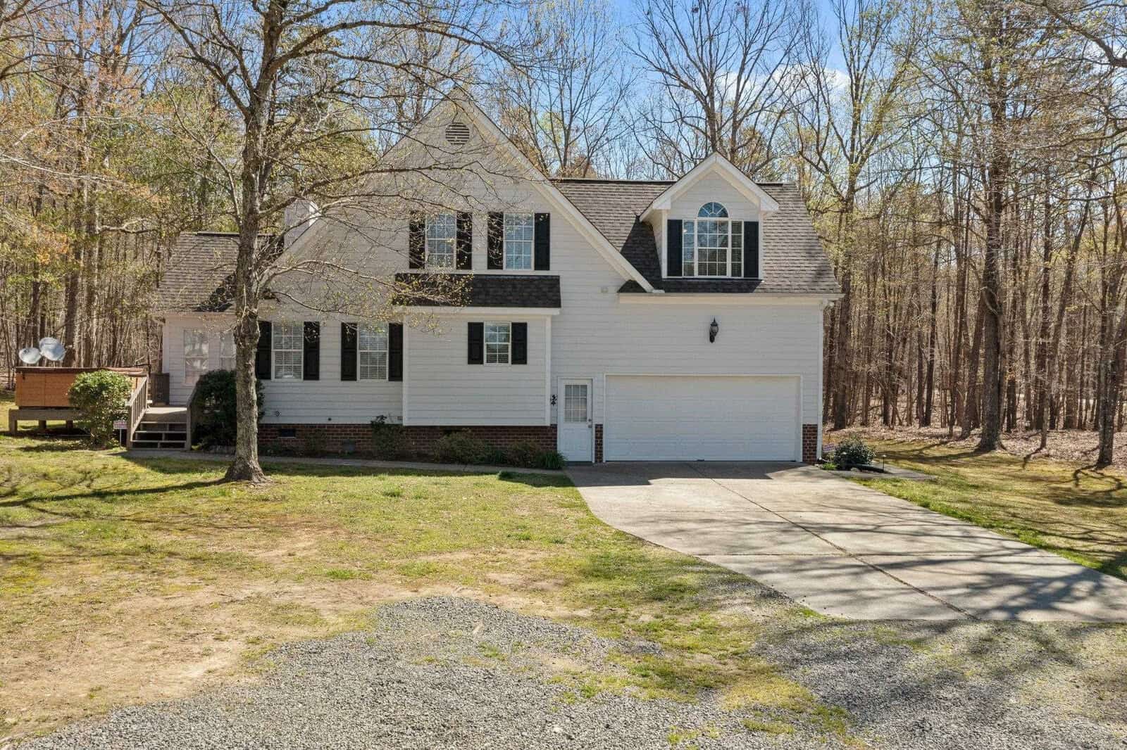 A two-story white house with dark shutters and a gabled roof is surrounded by trees. It features a driveway leading to a double garage and a front lawn with scattered trees and patches of grass, set against a backdrop of a wooded area under a clear sky.