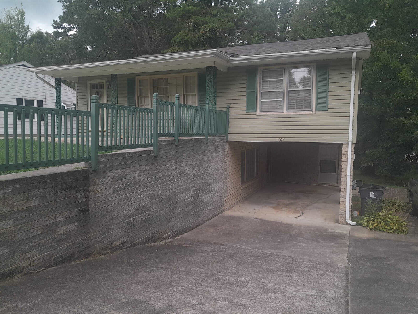 A single-story house with green siding, featuring a raised front porch with green railings, a stone retaining wall, and an under-house garage.