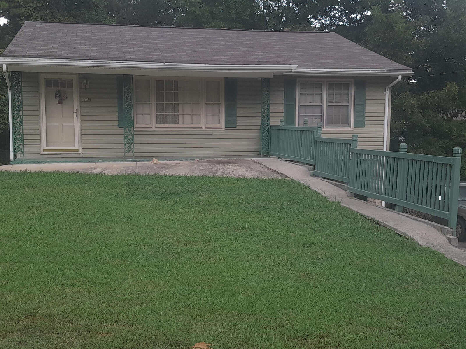 A single-story house with a green exterior and shutters, featuring a front porch with green railing, on a property with a well-maintained lawn.