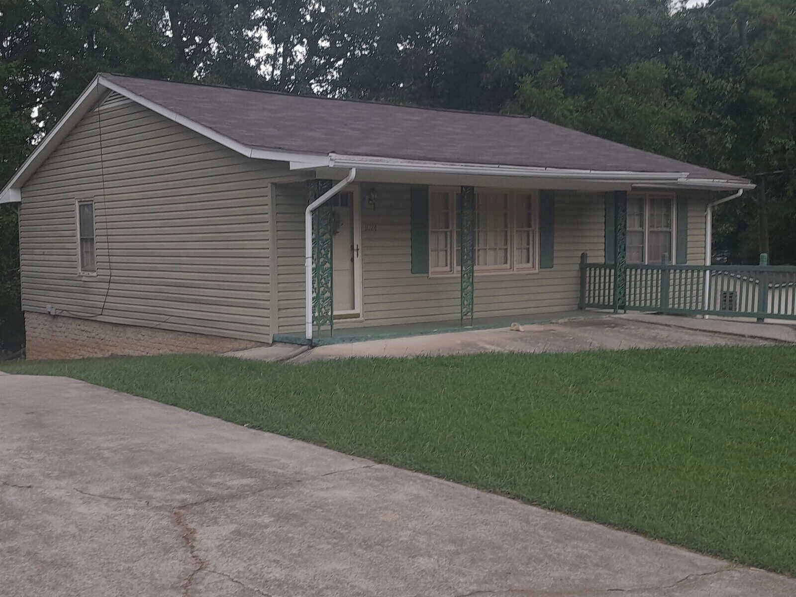 A single-story house with a covered front porch, grey siding, and a green roof. there is a concrete driveway on the left and a grass lawn in the foreground.