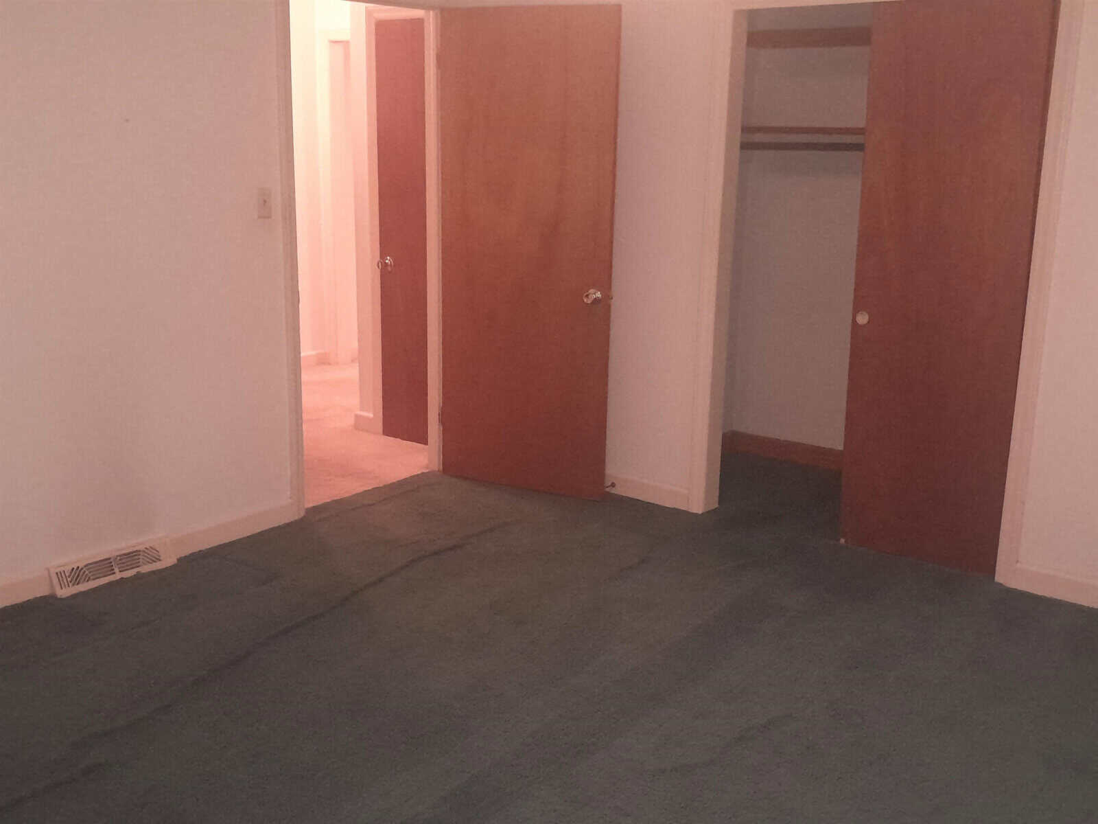An empty room with a grey carpet, two open wooden doors, and one closed door, with visible wrinkles in the carpeting near the center of the room.