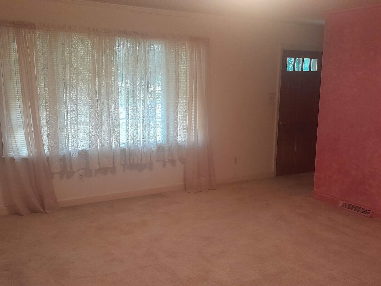 Empty room with beige carpeting, sheer curtains on a window, and a red accent wall to the right.