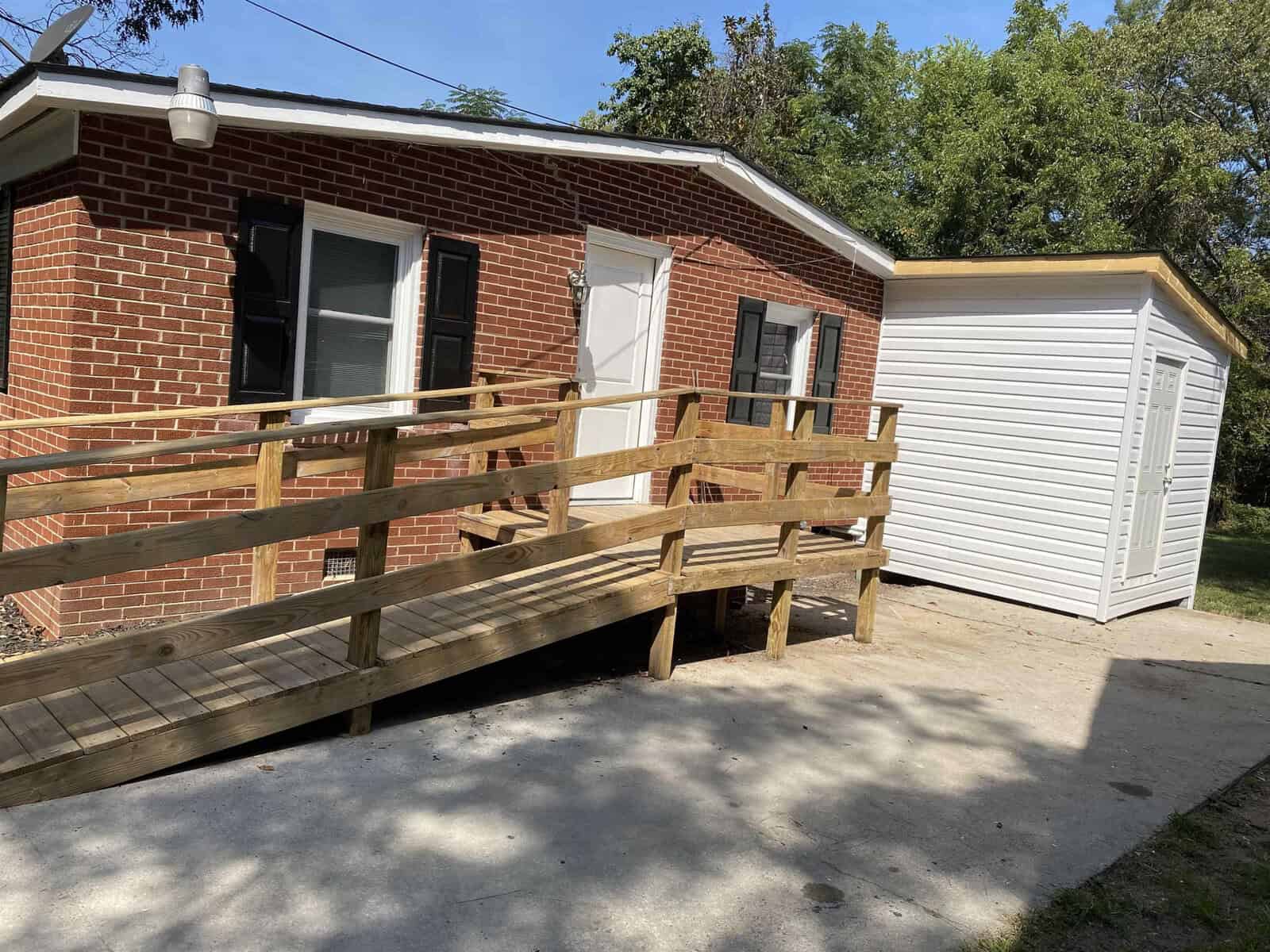 A single-story brick house with a wooden ramp leading to the front door and a small white storage shed located on the right side of the house.