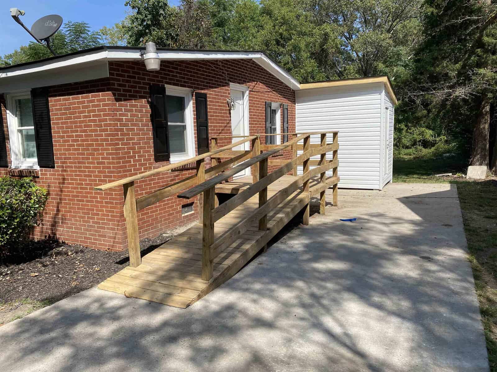 A small residential building with a brick exterior and a new wooden ramp leading up to the side entrance, with a white storage shed in the background and a concrete driveway in the foreground.