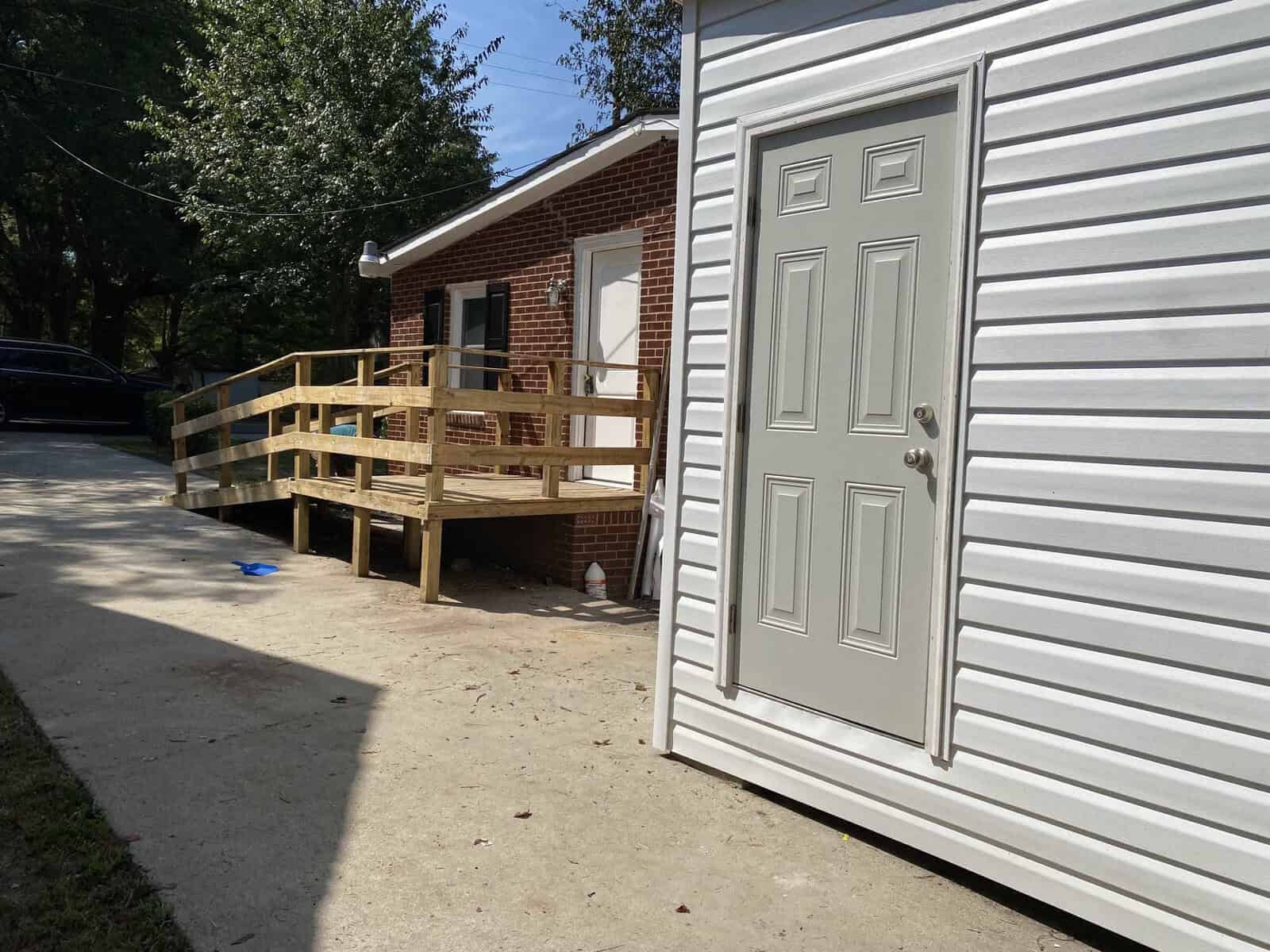A gray sliding door partially open, leading to a wooden deck attached to a brick building with trees in the background on a sunny day.