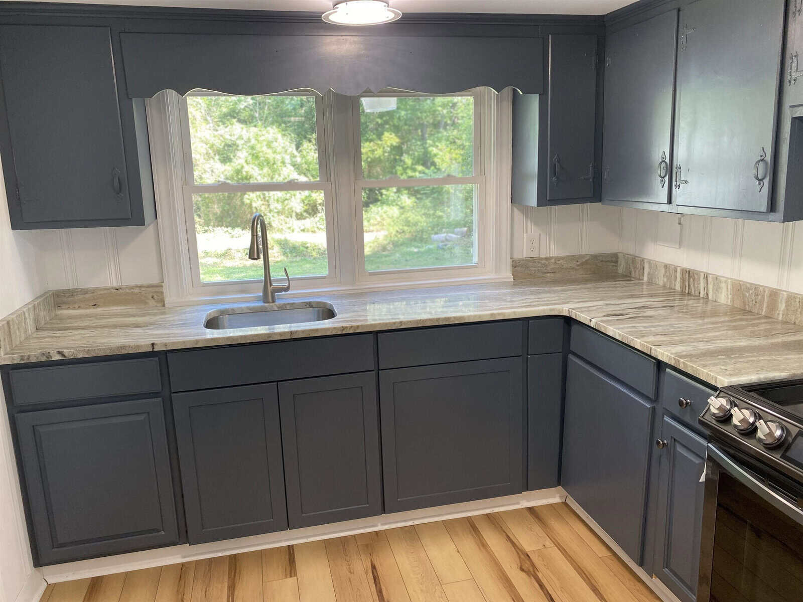 A small, modern kitchen with gray cabinets, stainless steel appliances, and a window above the sink overlooking a green outdoor area.