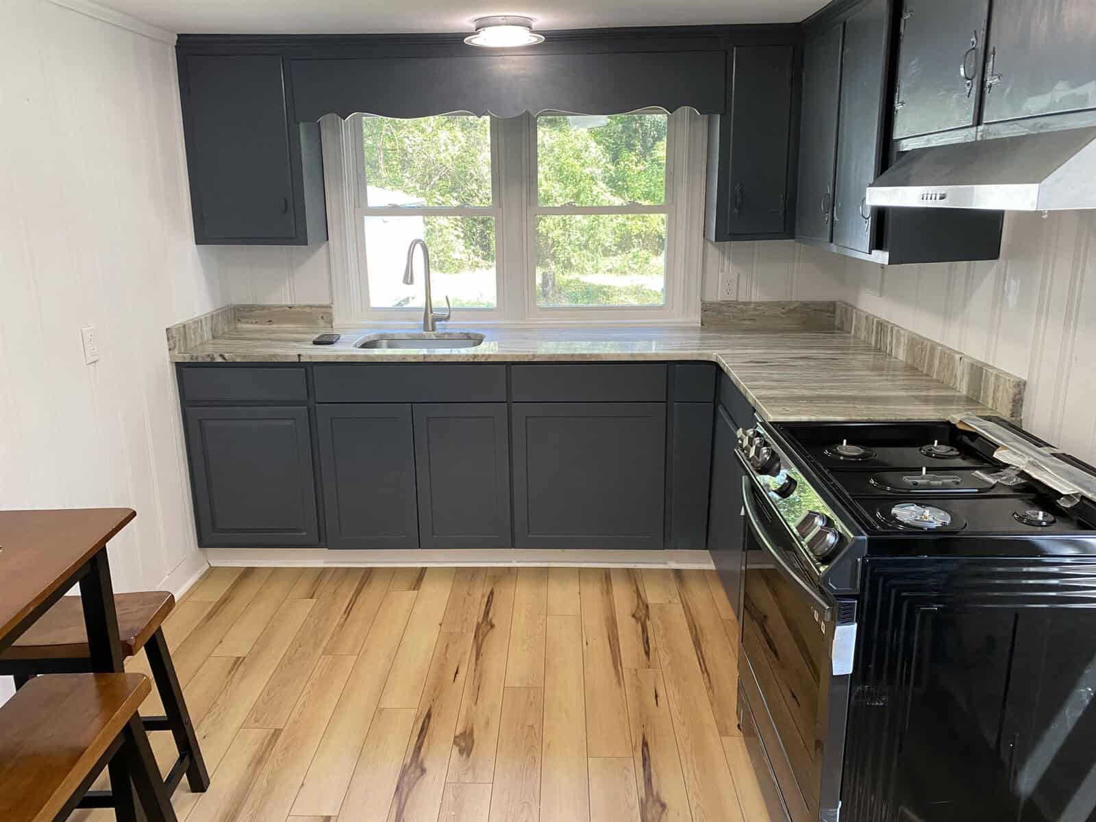 A compact kitchen with dark gray cabinets, a stainless steel stove, light wooden flooring, and a window above the sink.