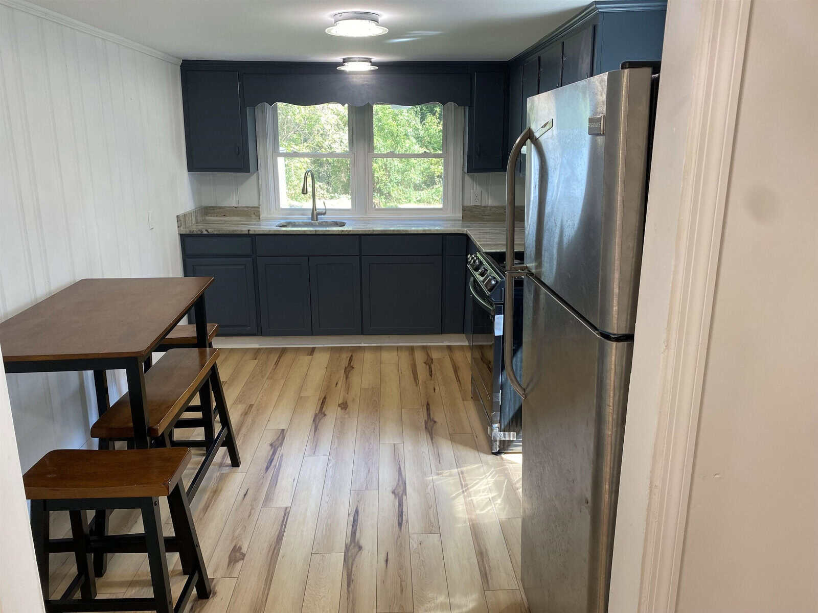 A small, modern kitchen with dark blue cabinets, stainless steel appliances, and a wooden dining table with stools.