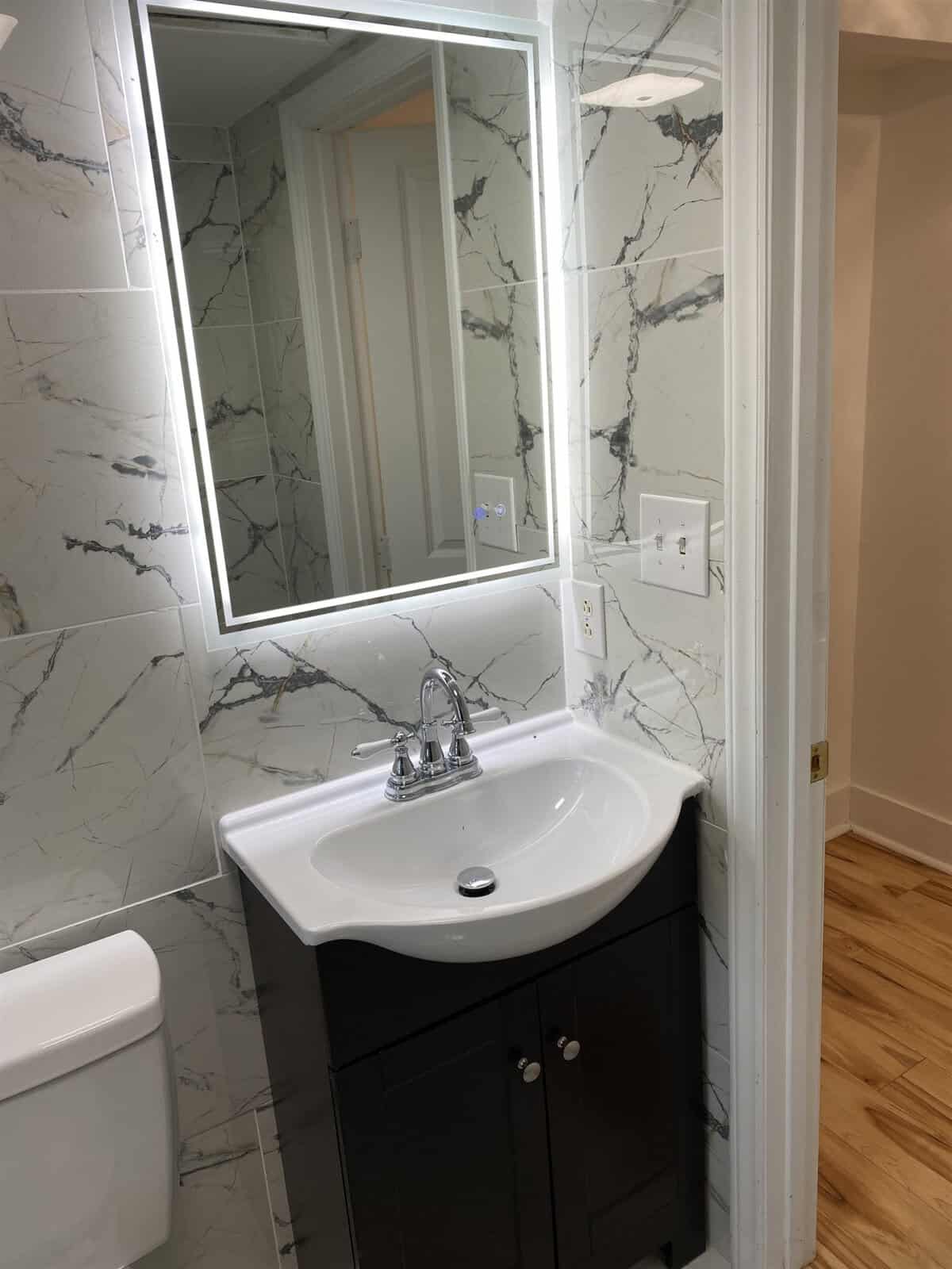 A modern bathroom featuring a white sink with a chrome faucet, mounted on a dark brown vanity cabinet, flanked by a toilet and a mirror with a marble-patterned wall in the background.