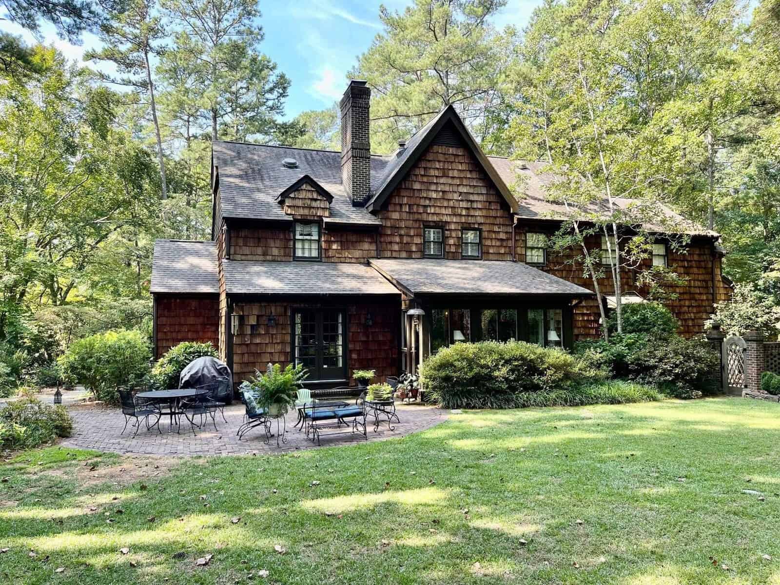 A quaint two-story wooden house with a shingled exterior, featuring a chimney, surrounded by lush greenery and a well-maintained lawn with an outdoor sitting area.