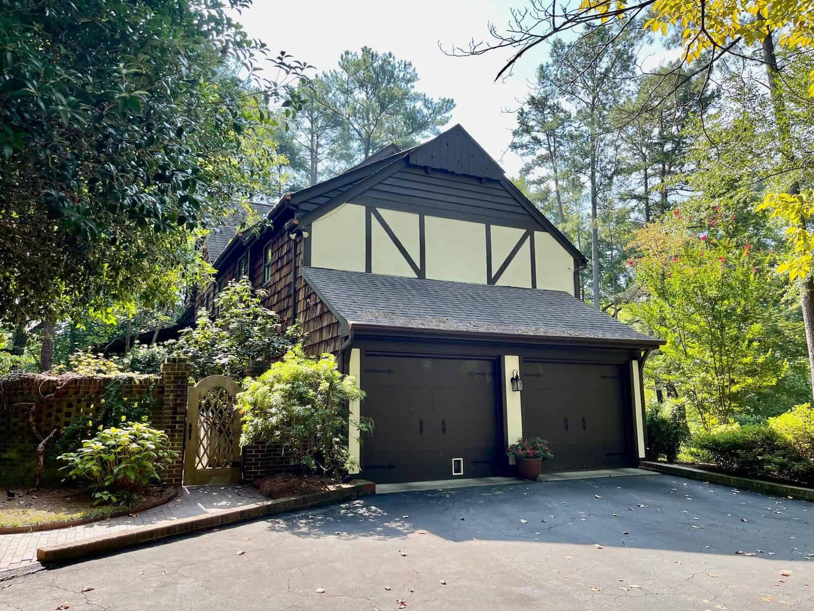 A two-story tudor-style house with a double garage, surrounded by trees and landscaping, under a clear sky.