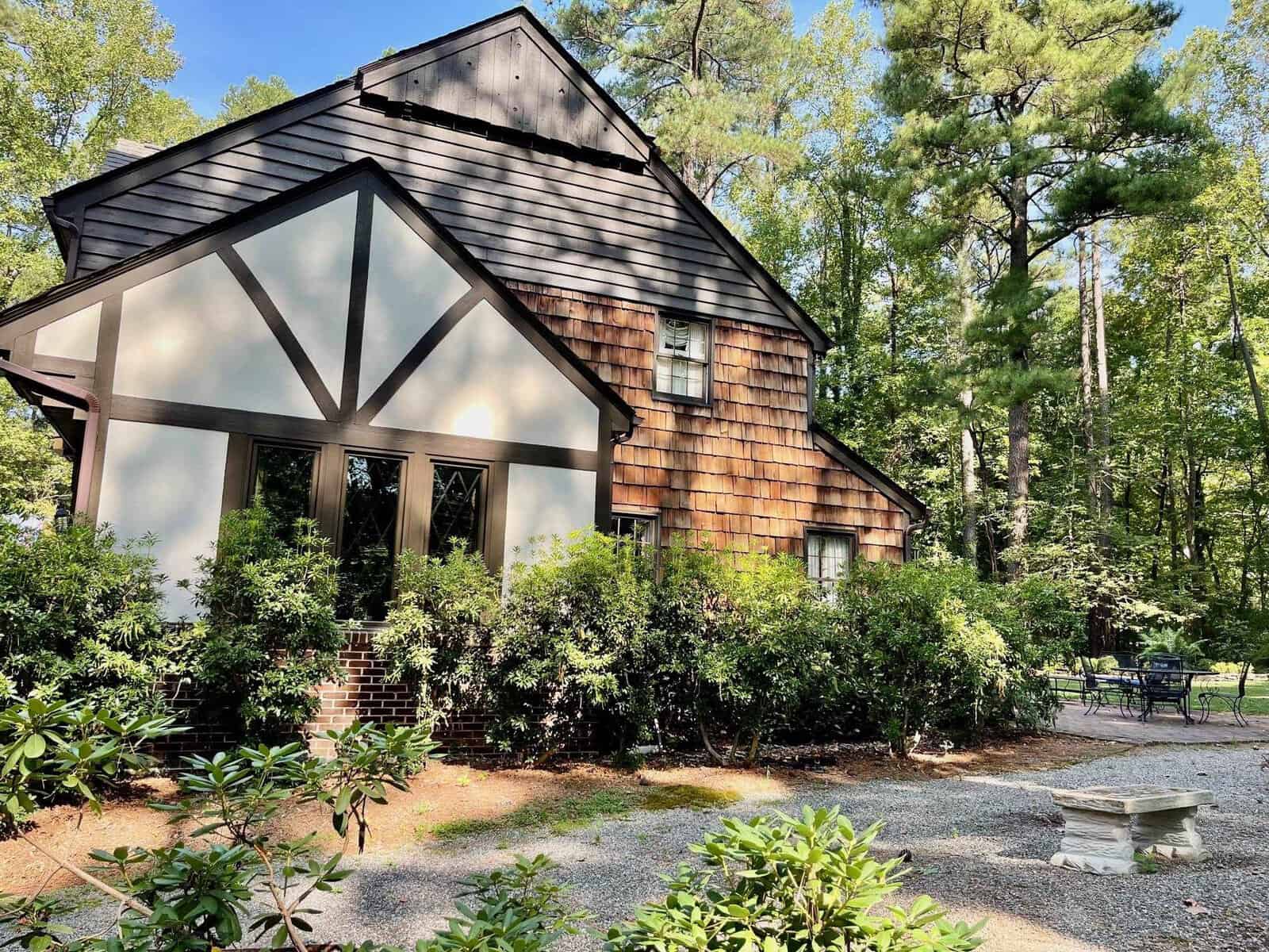 A two-story house with a combination of dark siding and wood shingles, featuring large windows and surrounded by lush greenery and a gravel path.