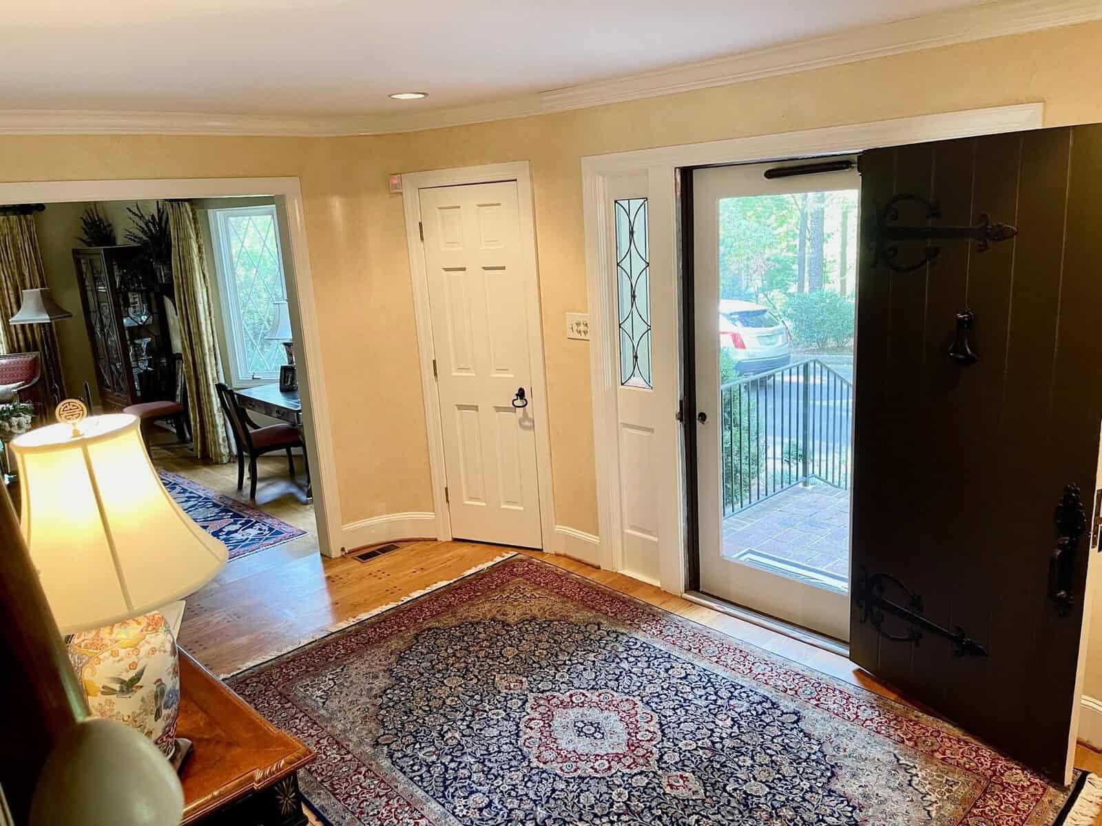 A well-lit entrance hall of a house featuring a decorative rug, a side table with a lamp, an open front door with glass side panels, and a closed white door to another room.