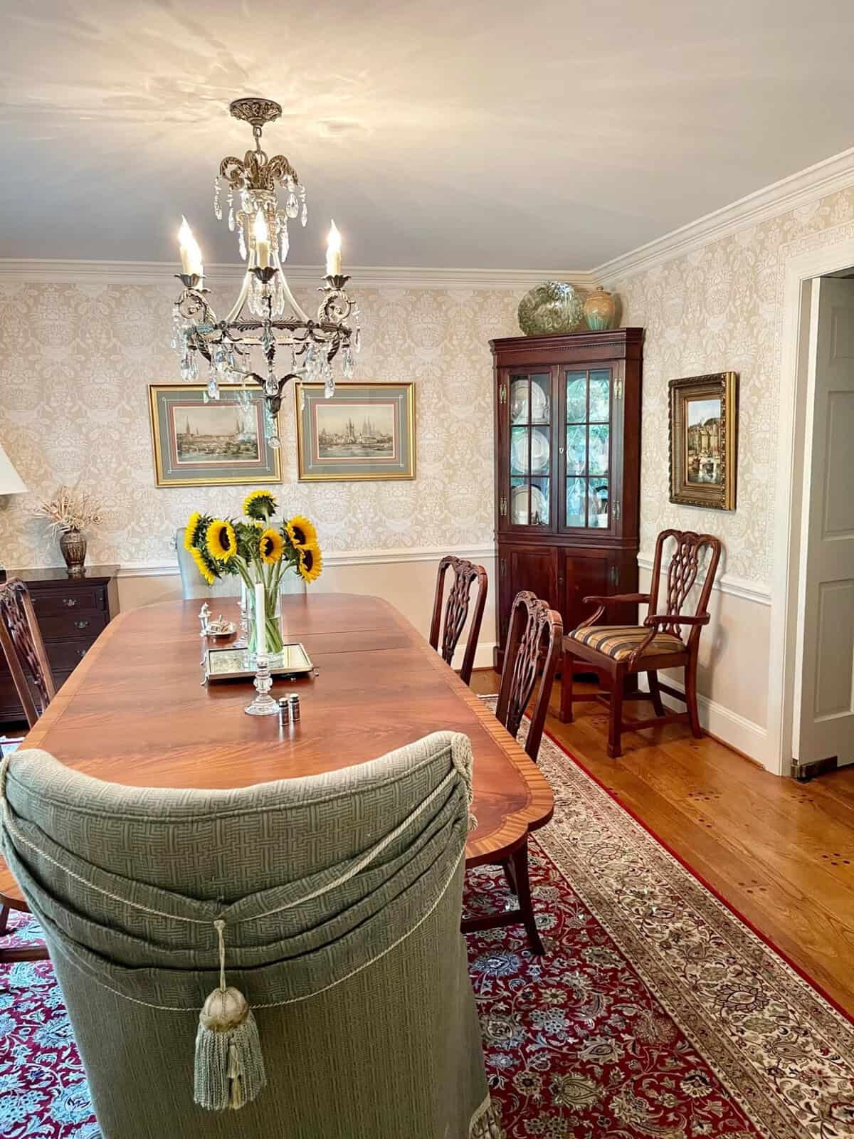 A traditional dining room with an elongated wooden table, upholstered chairs, an ornate chandelier, patterned wallpaper, and framed artwork on the walls.