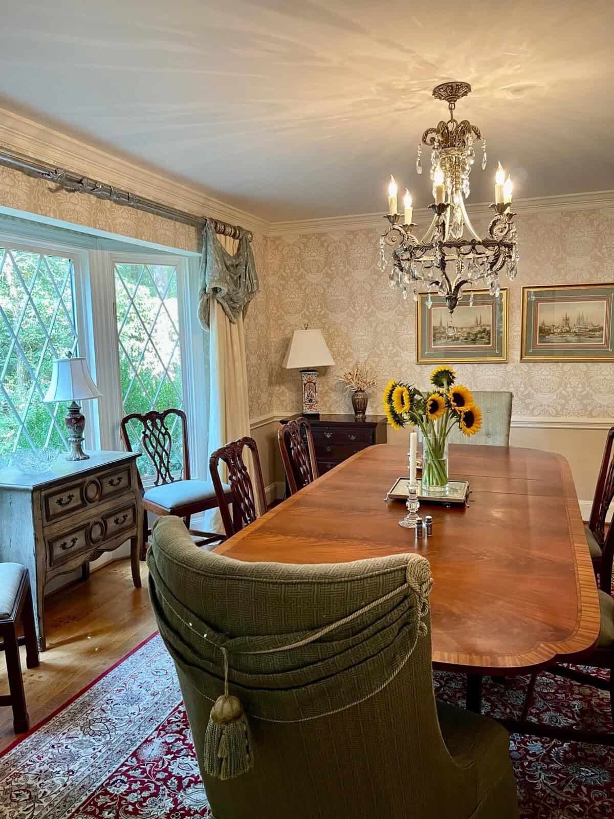 A traditional dining room featuring a large wooden table, upholstered chairs, and a chandelier, with a bouquet of sunflowers as a centerpiece.