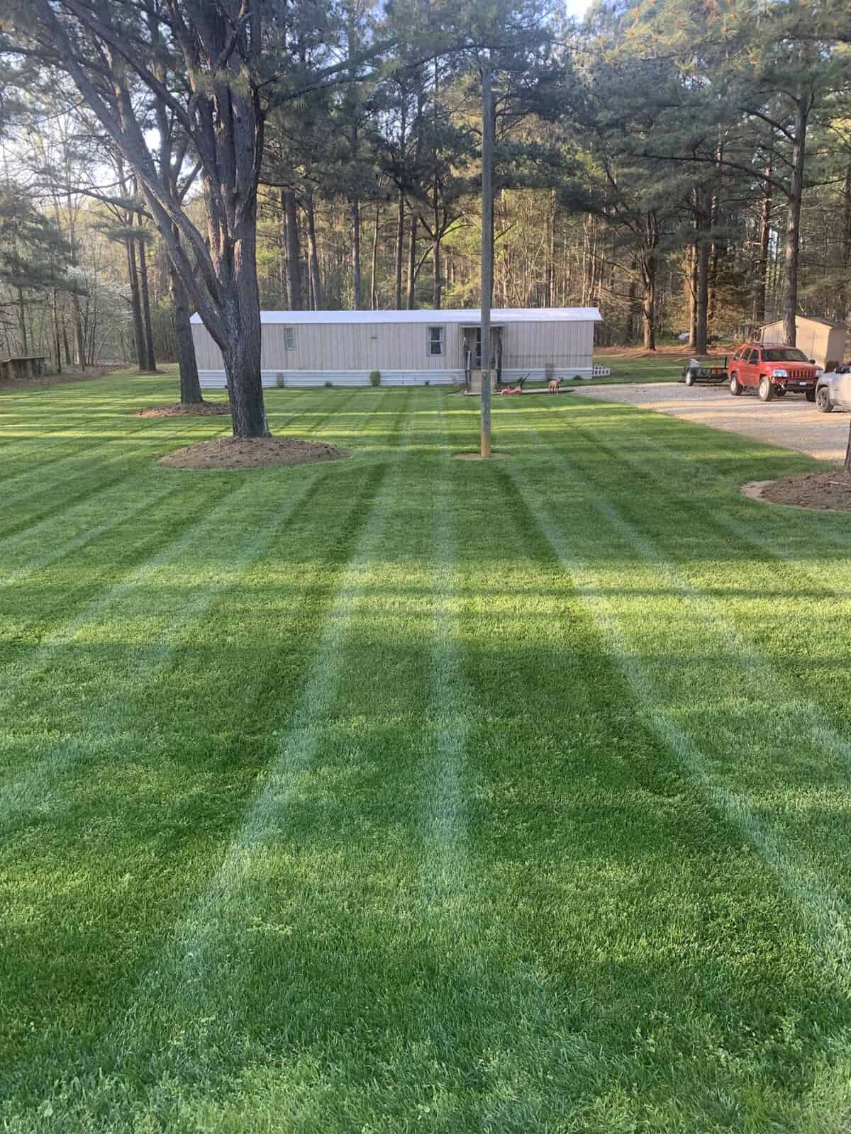 A neatly mowed lawn with striped patterns in front of a modern white rectangular building, surrounded by trees. The yard is bordered by a gravel driveway, with a red vehicle parked nearby.