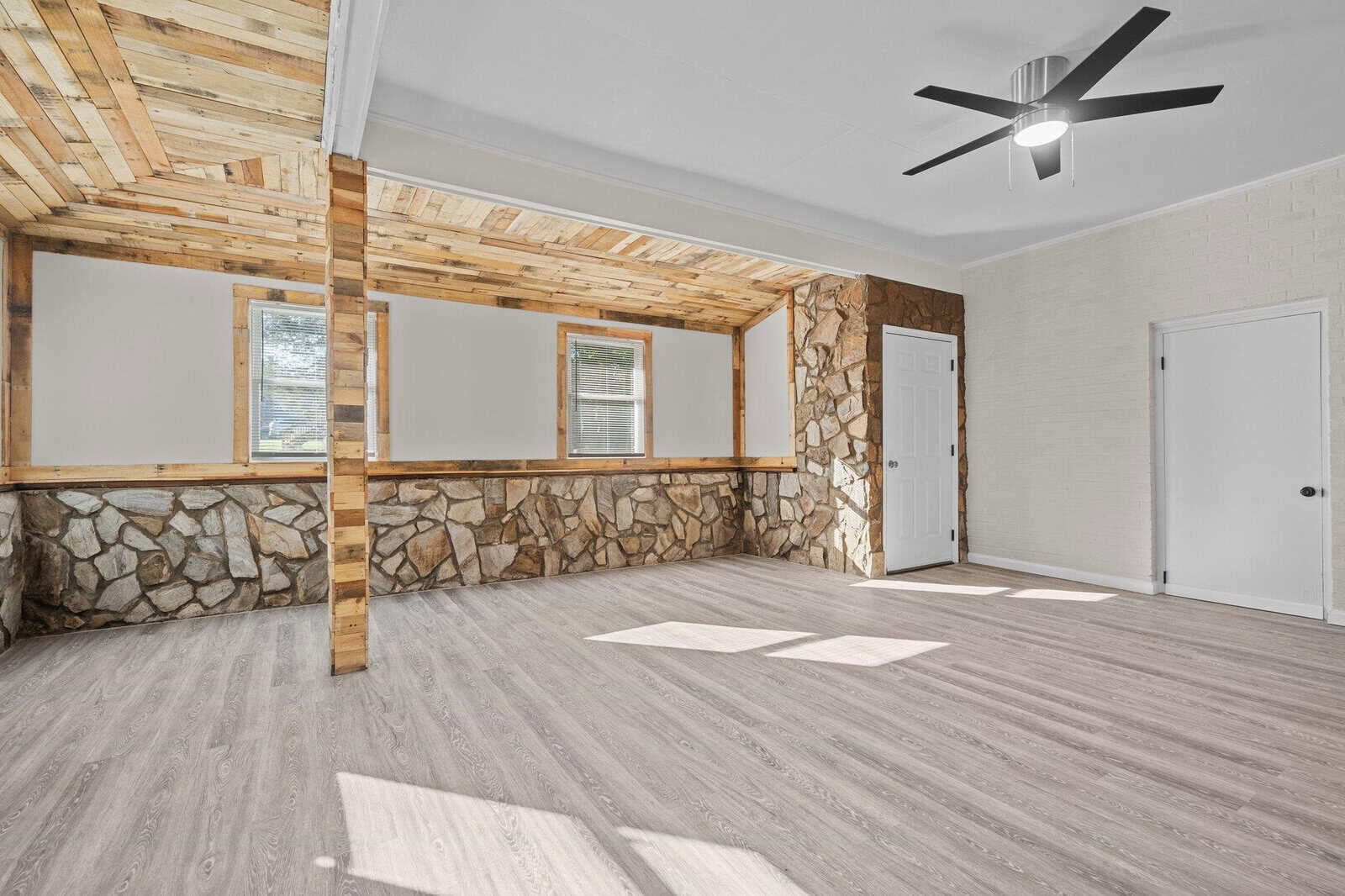 A spacious room with light gray flooring, white walls, and a wooden cathedral ceiling featuring a central supporting beam. the room is equipped with a modern ceiling fan, natural stone accent walls under the windows, and two white doors. sunlight filters through the windows, casting patterns on the floor.