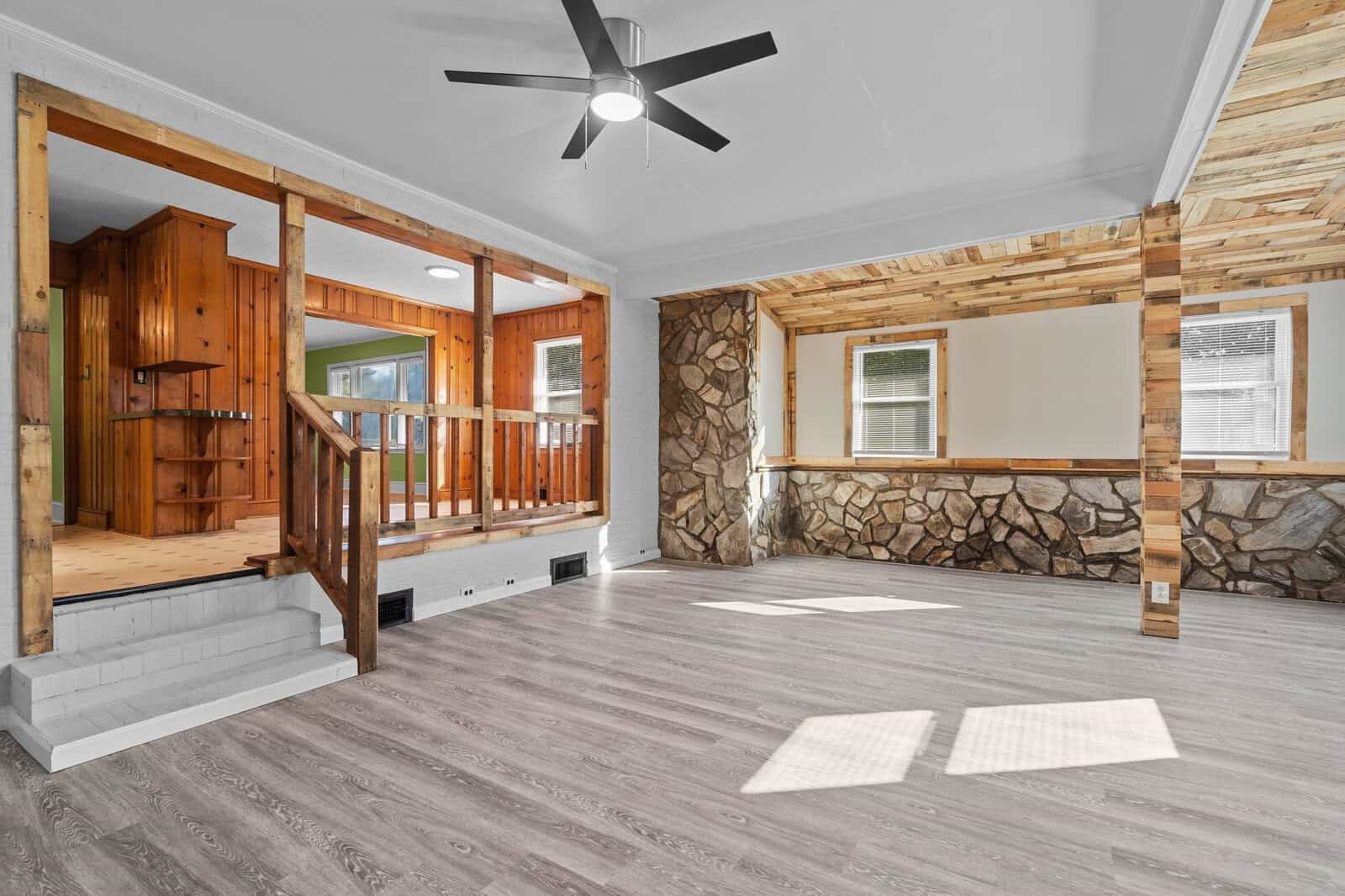 An empty room with wooden and stone wall accents, featuring a ceiling fan, hardwood floors, and a wooden railing separating an elevated section.